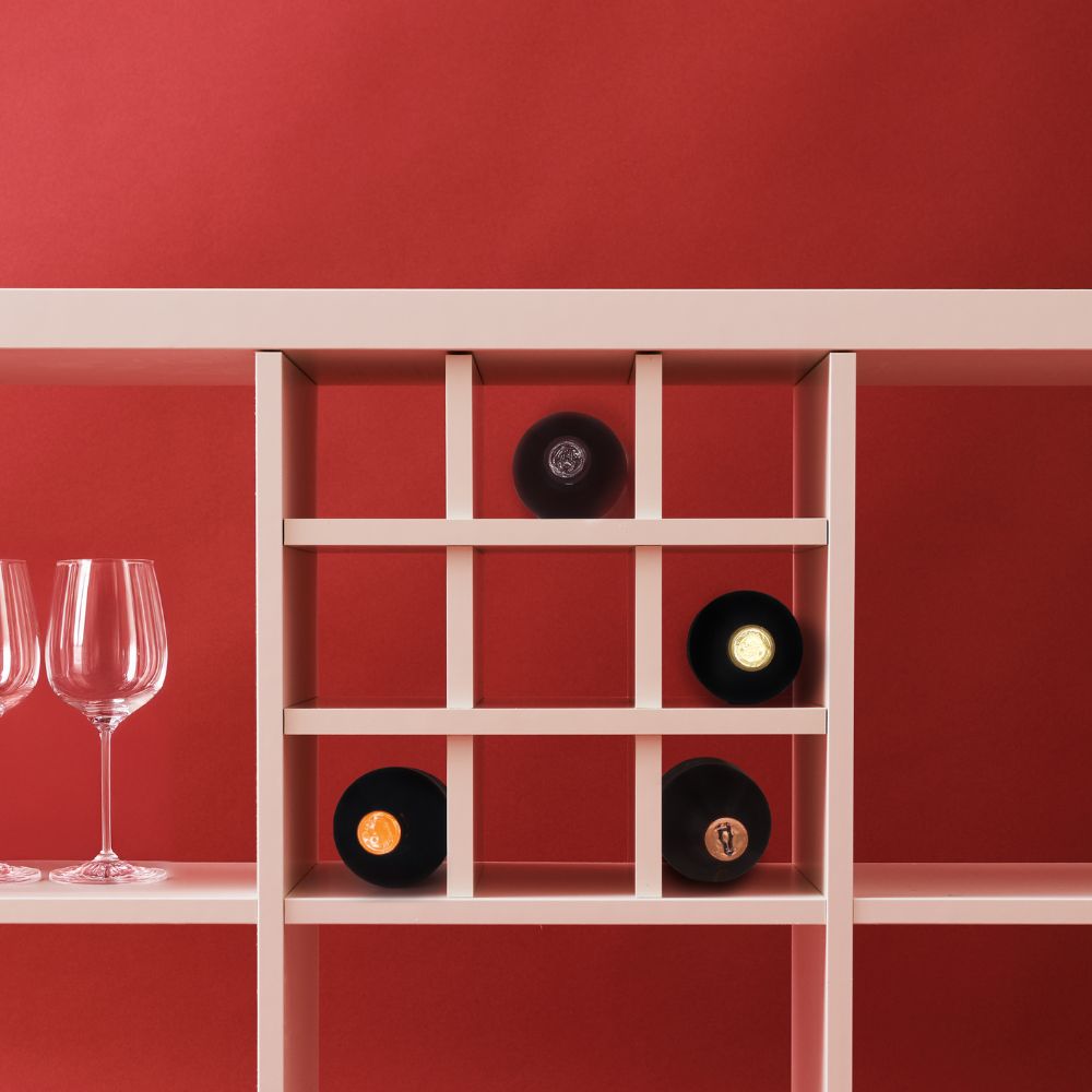 Ansicht eines Kallax Fachs vor rotem Hintergrund. In dem Fach ist ein Regaleinsatz, der das Kallax Fach in 9 kleine Fächer unterteilt. Dieser dient als Flascheneinsatz, 4 Weinflaschen werden hier liegend gelagert.