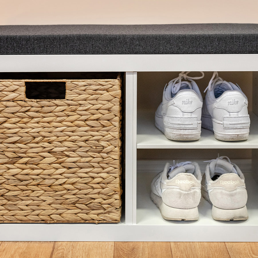 Ein weißes Kallax Regal mit Schuhen und einem Korb. Darauf liegt eine Sitzauflage in Anthrazit.