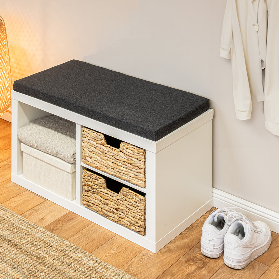Eine weiße IKEA Kallax Sitzbank mit einem Sitzpolster in Anthrazit. Die beiden Regalfächer sind mit Körben gefüllt.