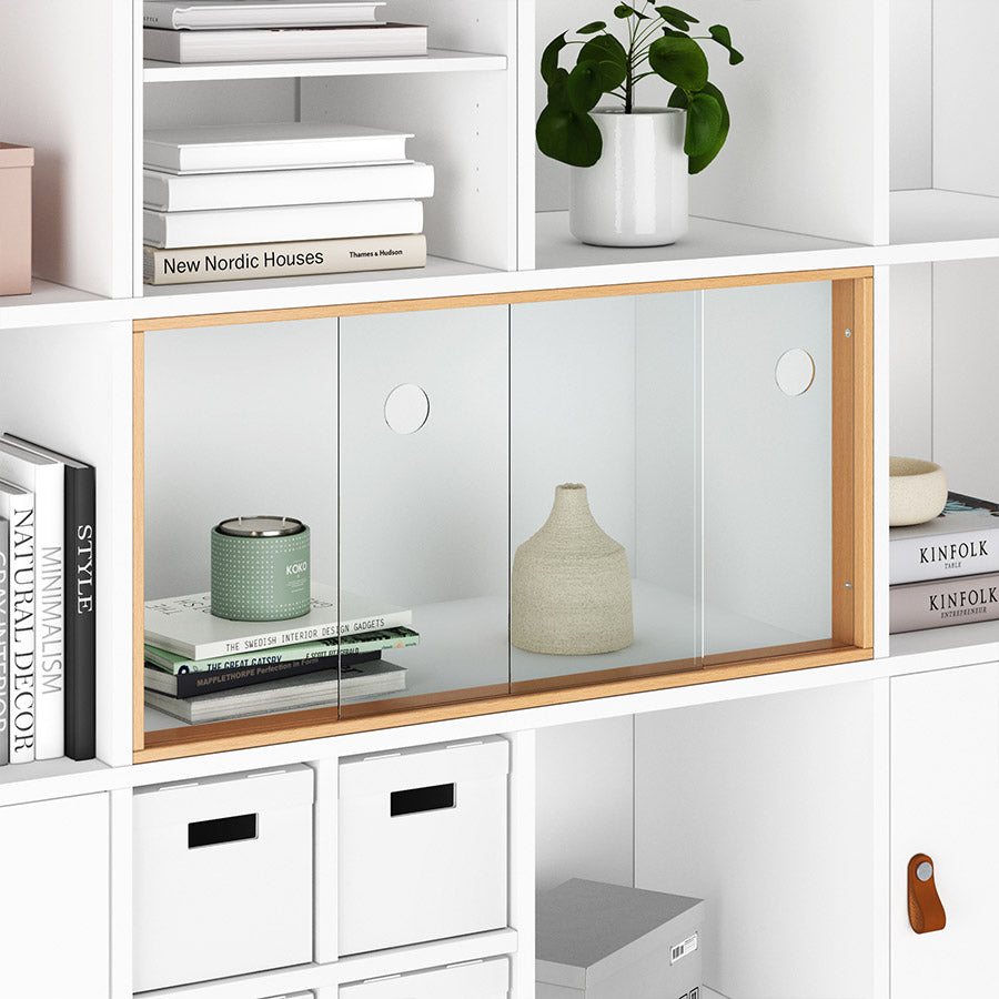 Weißes Kallax Regal mit Schiebetür aus Glas und verschiedenen Dekoobjekten, wie Vasen und Bücher.