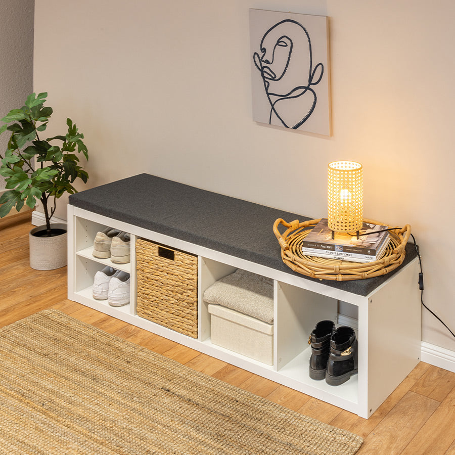 Ein weißes Ikea Kallax Regal mit 4 Fächern liegt als Sitzbank längs auf dem Boden. Auf der Sitzbank liegt eine Sitzauflage in anthrazit aus Baumwolle. Auf dem Sitzpolster sieht man noch Deko-Artikel.